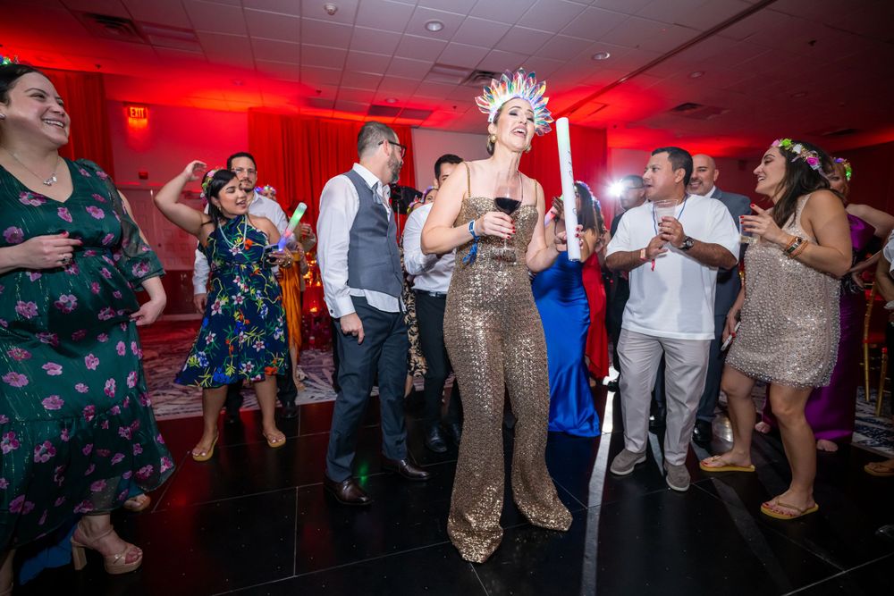 Wedding guests dance and celebrate around performer in sequined outfit under red lighting.