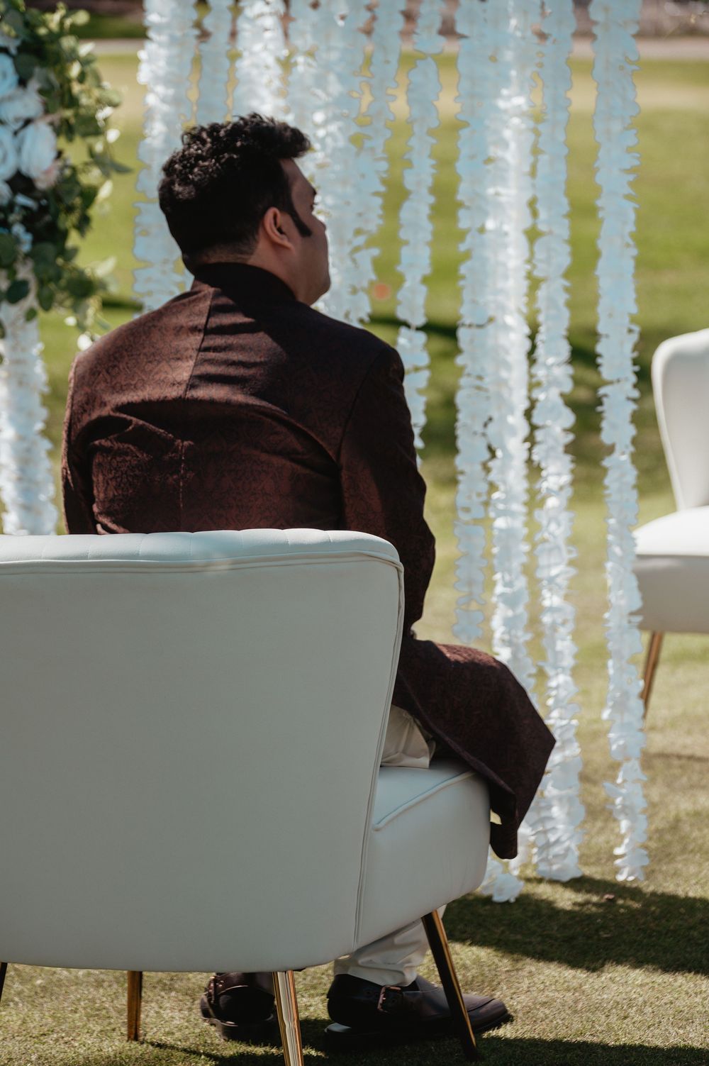 Groom in dark brown sherwani sitting on white chair at outdoor South Asian wedding ceremony.