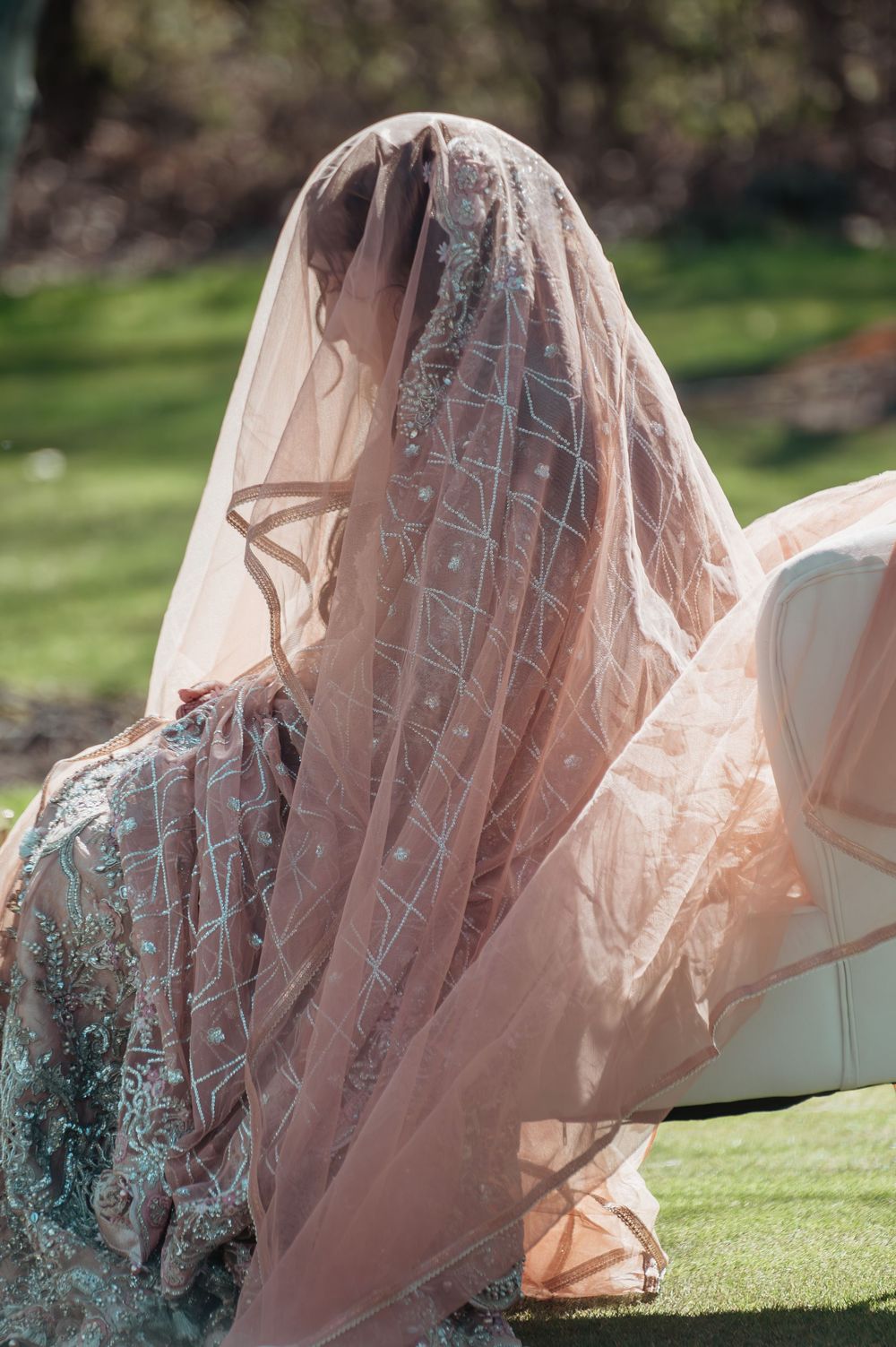 A bride wearing an ornate blush pink embroidered dupatta veil draped over her head, seated outdoors in natural light.