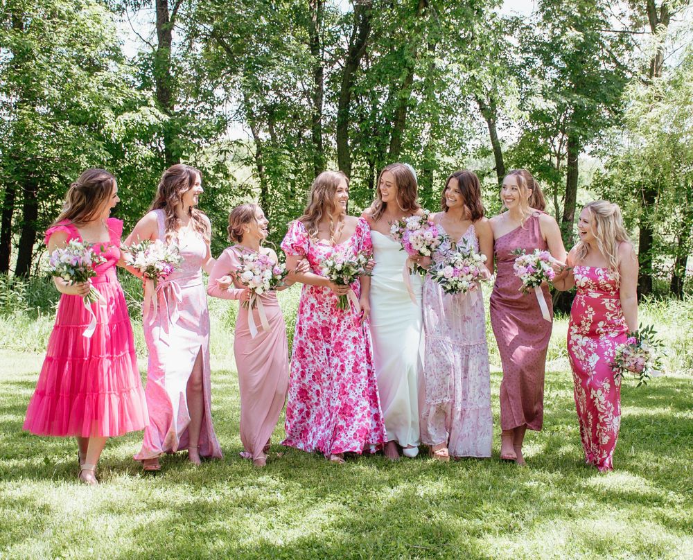 Wedding party in pink and floral dresses pose together on sunny lawn with bouquets.