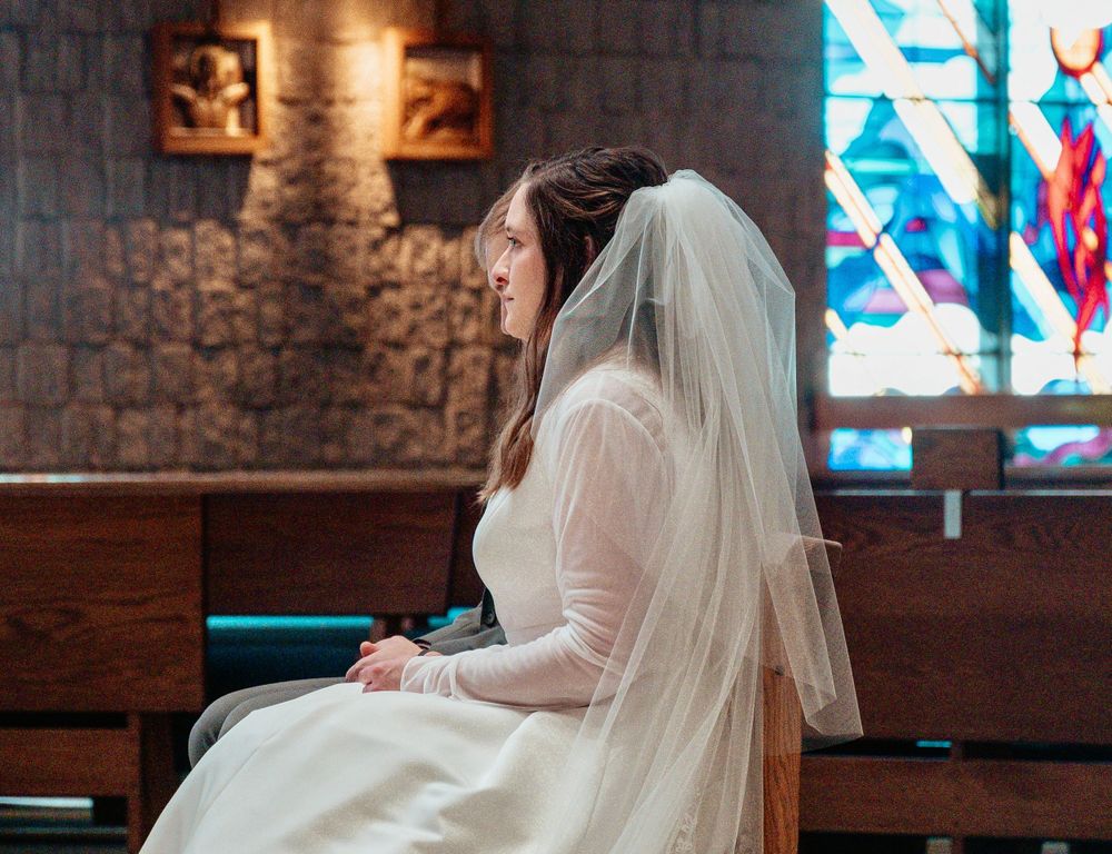 Side view of bride in white dress and veil sitting in church pew.