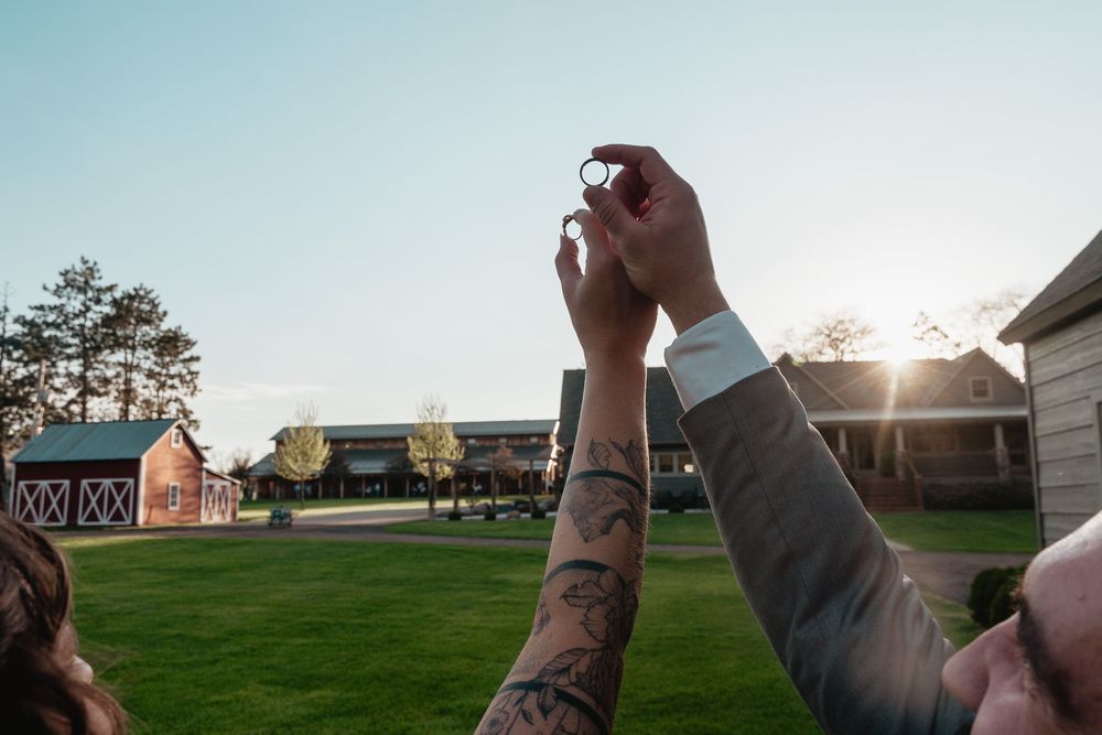 Raised hands with tattoos toasting in celebration against a sunset sky at an outdoor venue.