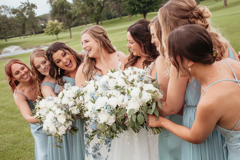 Group laughing together around white floral bouquet in light blue bridesmaid dresses.