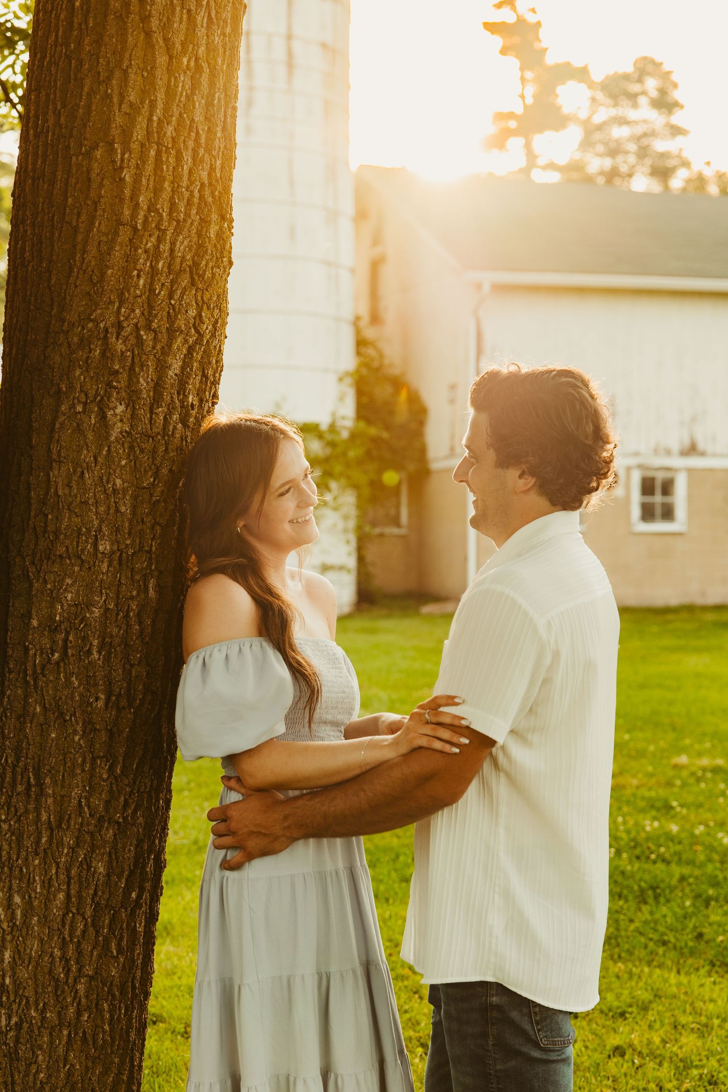 A romantic backlit couple embraces next to a tree at sunset with a white house in the background. Novi Northville Portrait Photographer