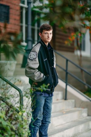 Student wearing varsity jacket and jeans poses on brick building steps with greenery.