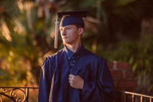 Person wearing navy graduation gown poses against sunset foliage.