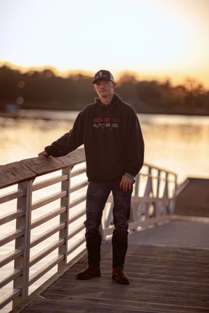 Person in dark hoodie leans on wooden dock railing at sunset.