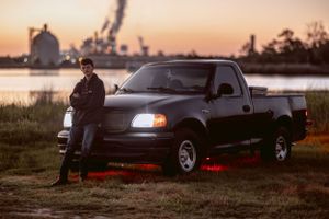 Dark pickup truck parked by waterfront with industrial background at sunset.