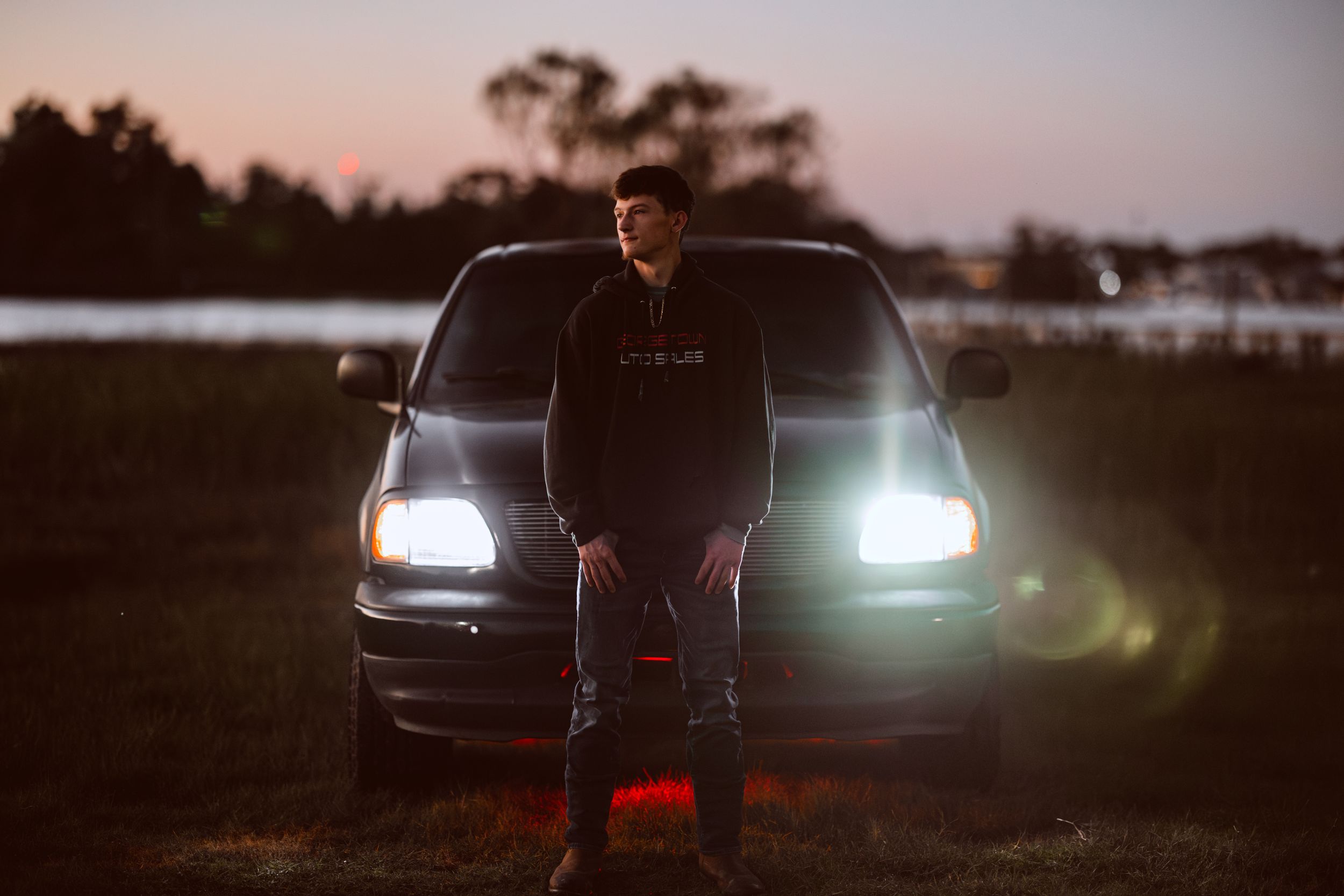 Silhouetted figure stands in front of car with illuminated headlights at dusk in outdoor setting.