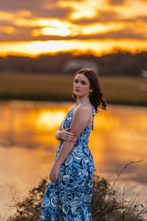 Woman in blue patterned dress poses by water during dramatic orange sunset.