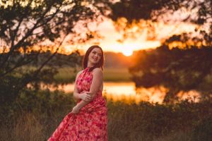 Woman in flowing red dress poses against golden sunset lake backdrop.
