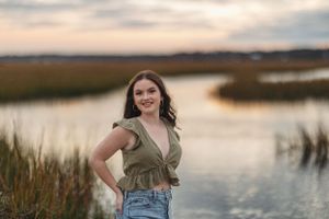 Person in ruffled green top and jeans poses by marsh at dusk.