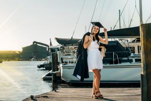 Graduate in white dress and graduation cap poses on dock with boats in background at sunset.