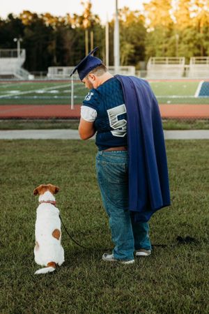 Graduate in blue cap and gown stands with dog on athletic field at sunset.