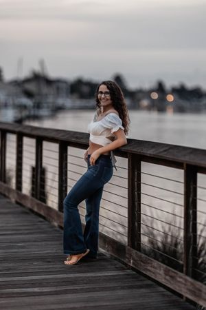 Portrait of woman in white crop top and jeans posing on waterfront boardwalk at dusk.