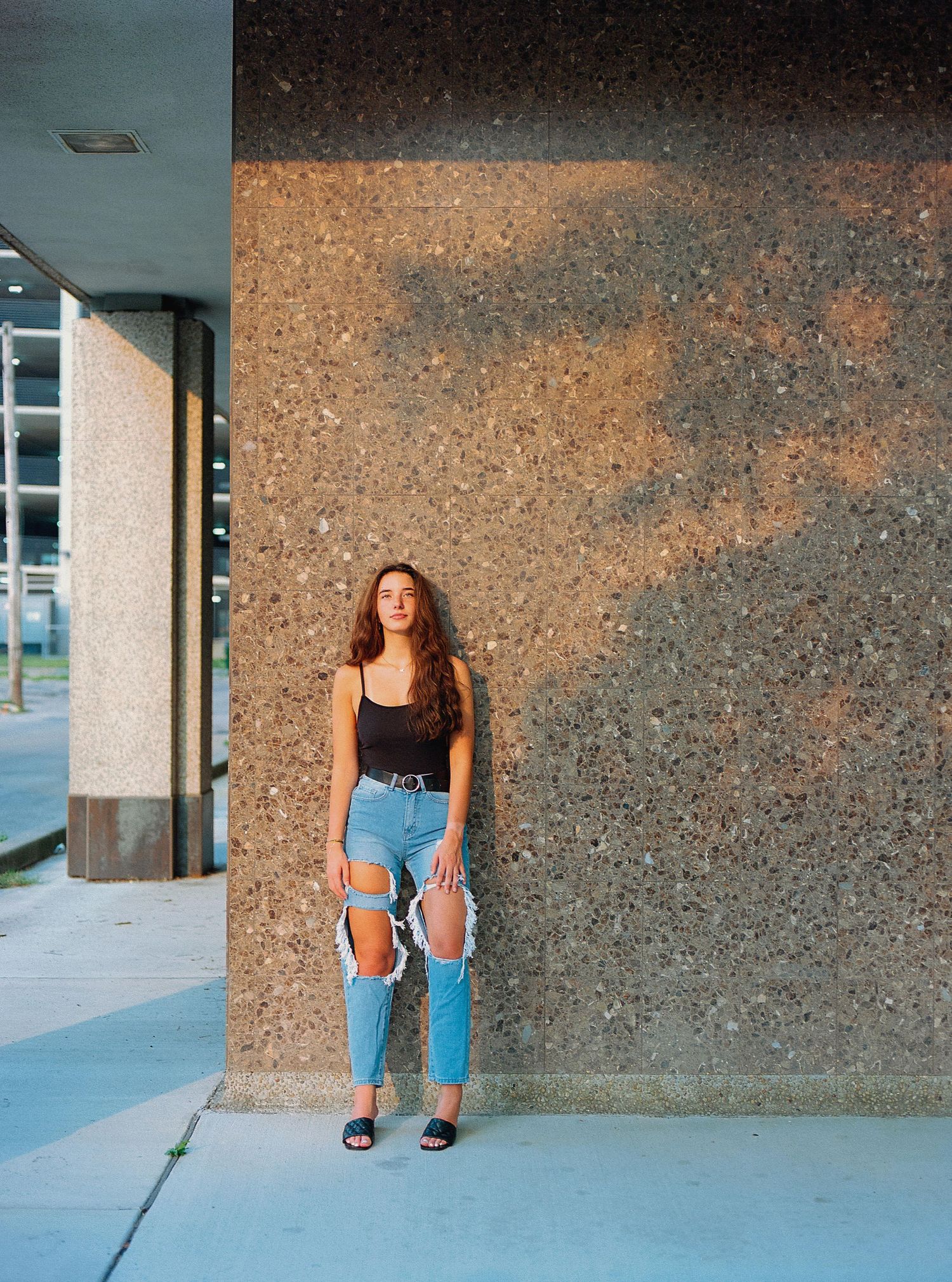 A casual fashion pose against a textured wall wearing distressed jeans and a black tank top.