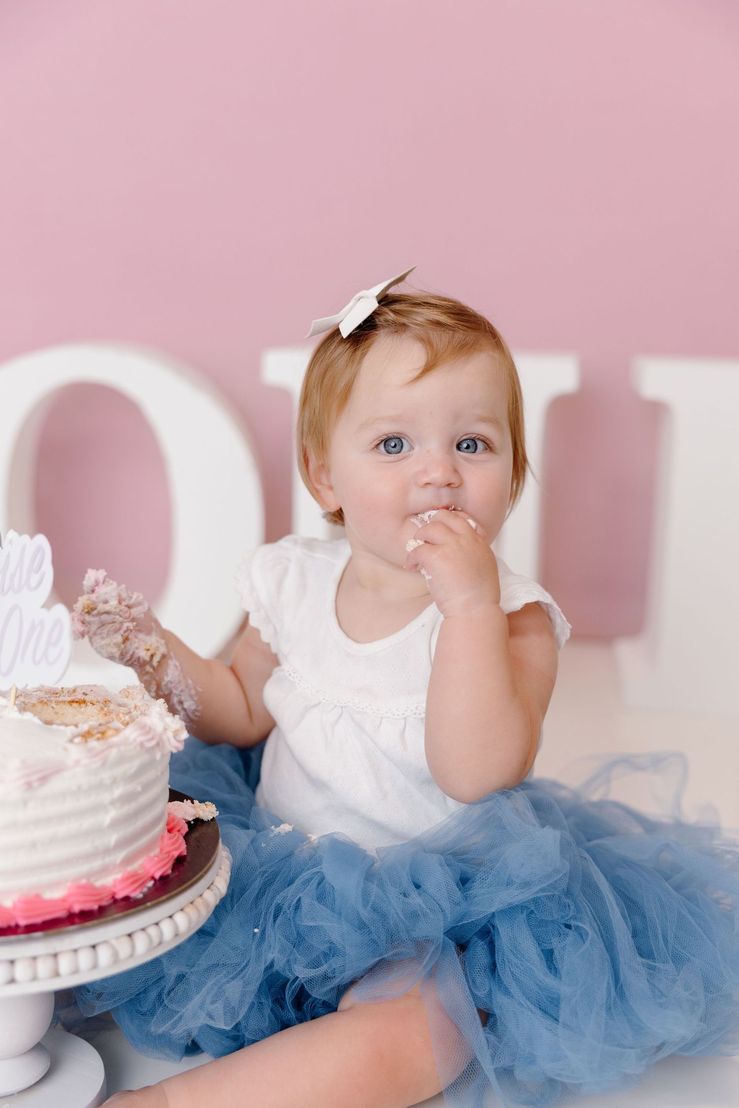 Baby in blue tutu and white top sits near pink birthday cake in series of cake smash photos.