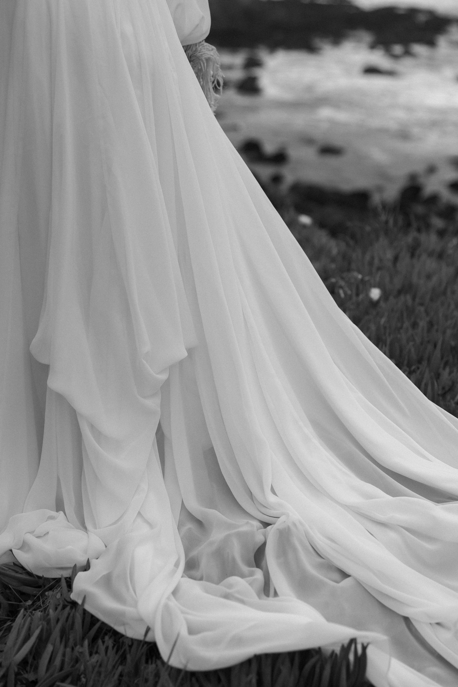 A flowing white wedding dress train draped elegantly across grass in black and white photography.