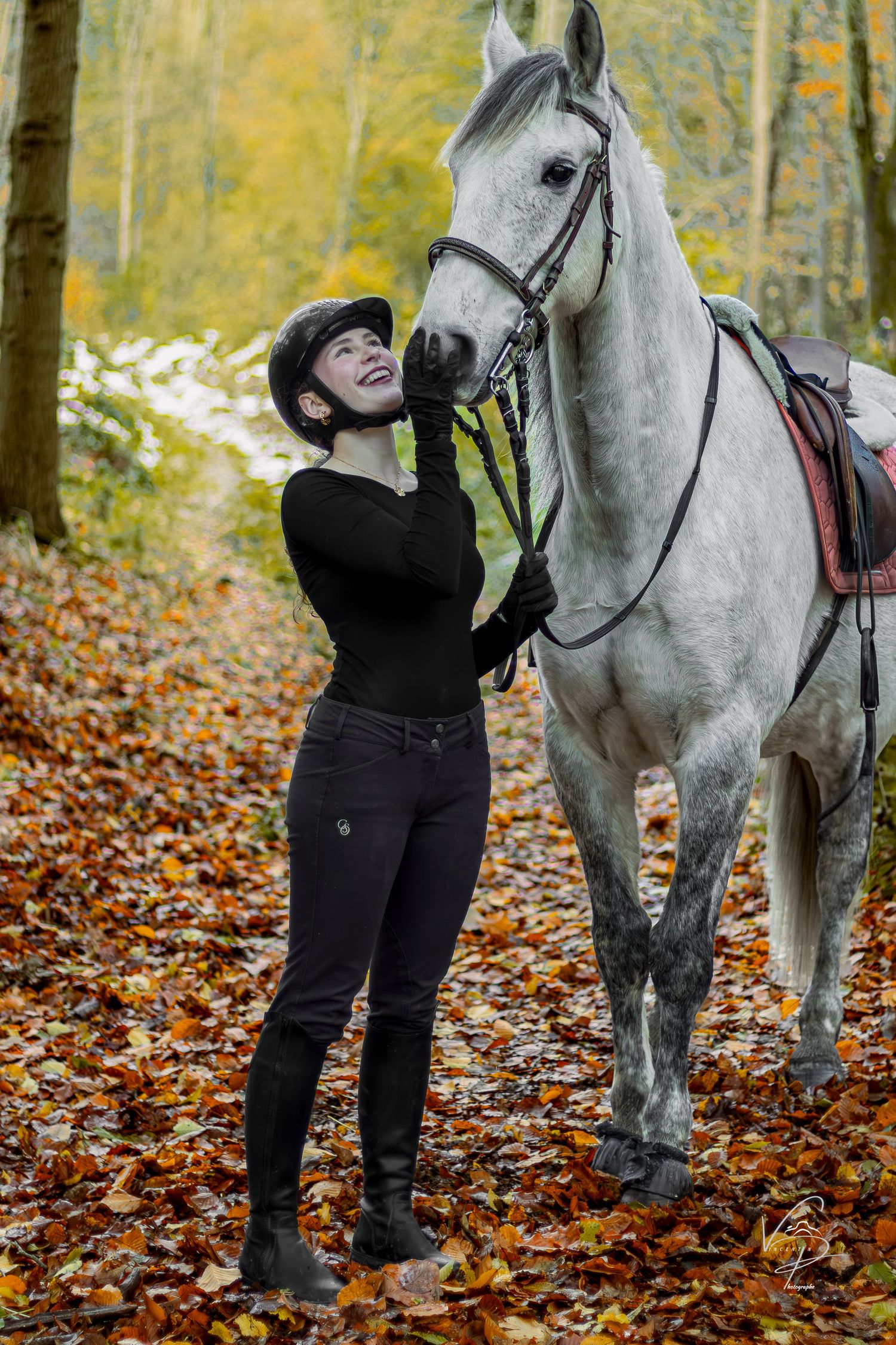 A person in black clothing interacts with a white horse in an autumn forest setting with fallen leaves on the ground.