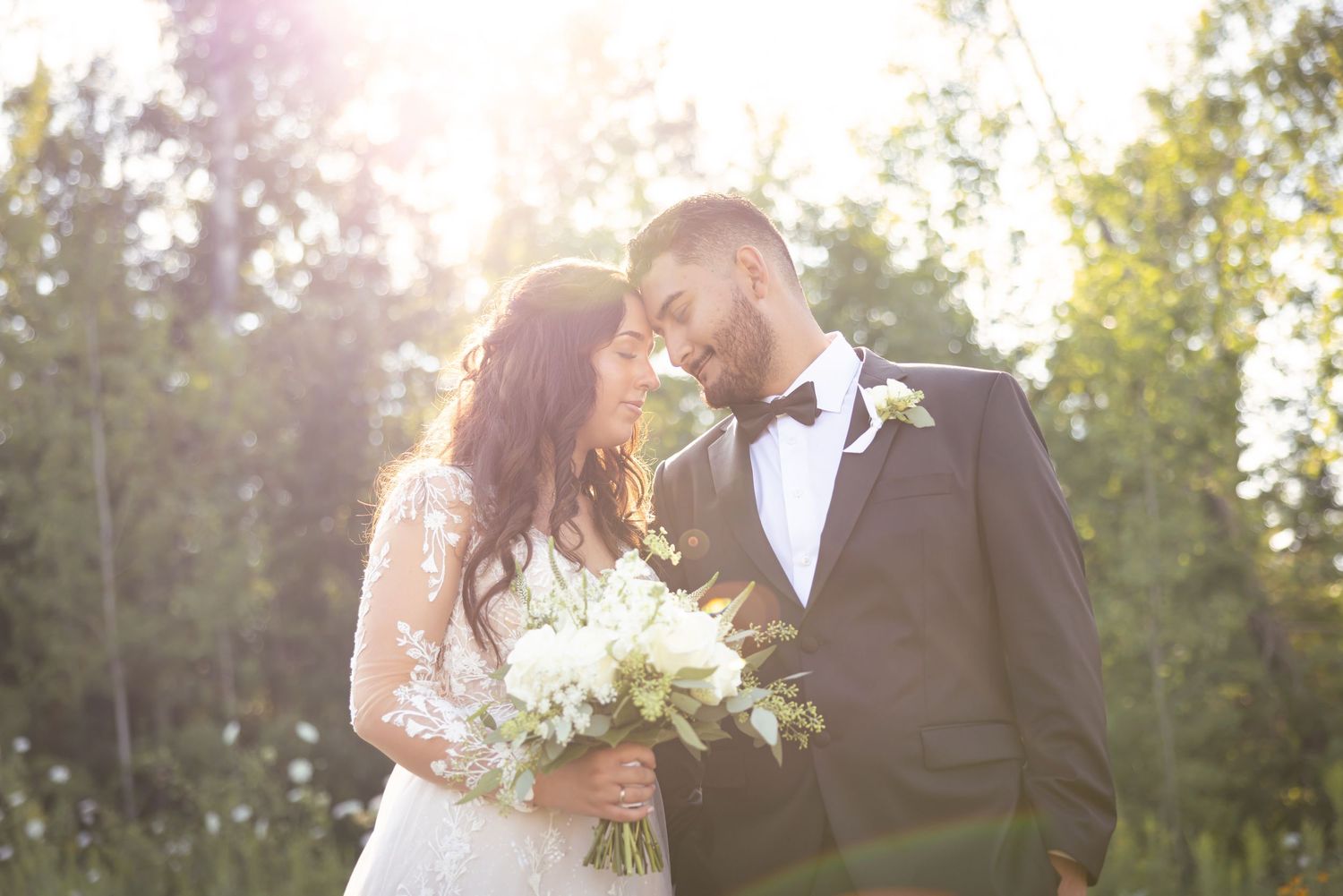 Newlyweds share a romantic kiss at sunset while holding a vibrant flower bouquet.