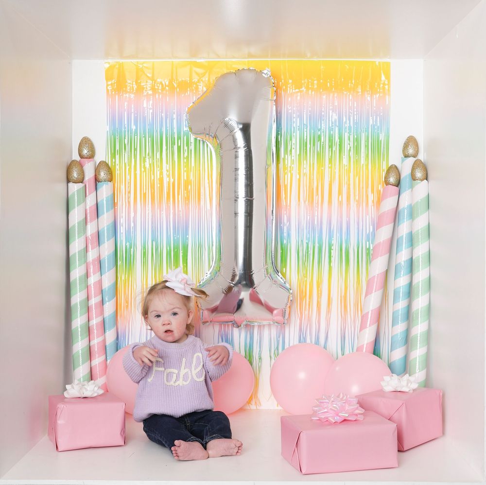 A baby's first birthday cake smash photo session with pink decor, rainbow backdrop, and pastel party decorations.