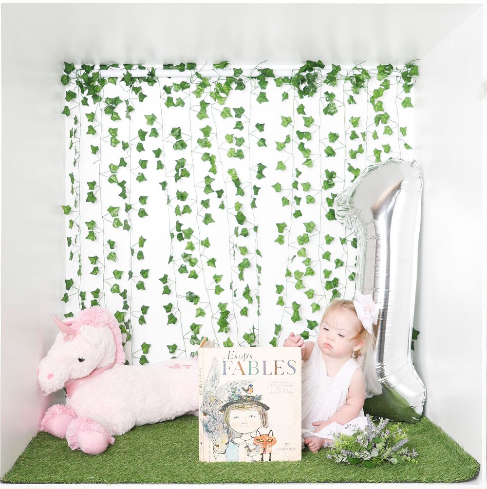 Baby photography set with green ivy wall backdrop, white furniture, and pink plush decorations.