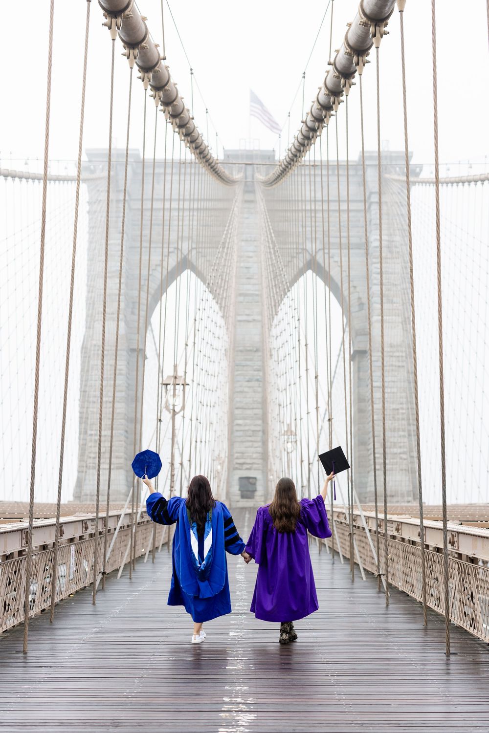 Two graduates in blue and purple academic regalia walking along the misty Brooklyn Bridge boardwalk.
