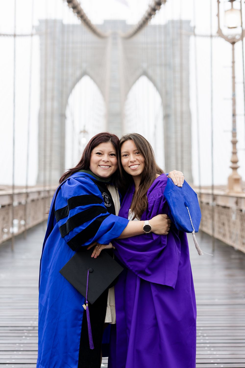 Two graduates in blue and purple gowns embrace while celebrating on the Brooklyn Bridge.