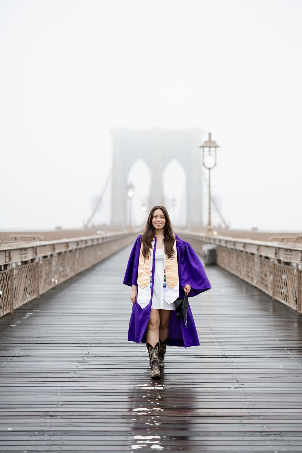 Graduate in purple academic regalia poses for photos on the foggy Brooklyn Bridge pedestrian path.
