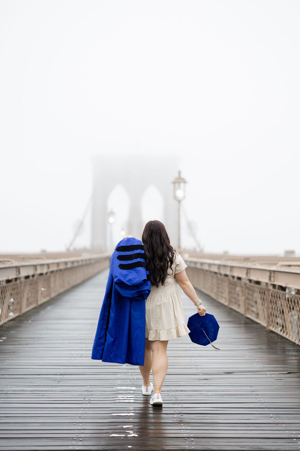 Graduates in blue and white outfits walk along the Brooklyn Bridge on a foggy day.