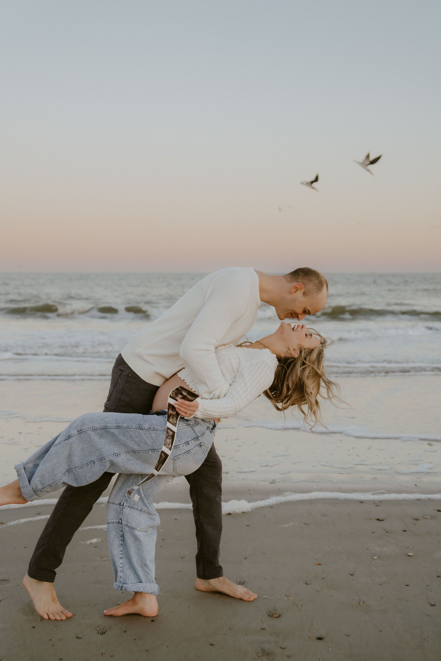 A couple in white attire walks along a sandy beach path at sunset with palm trees in the background.