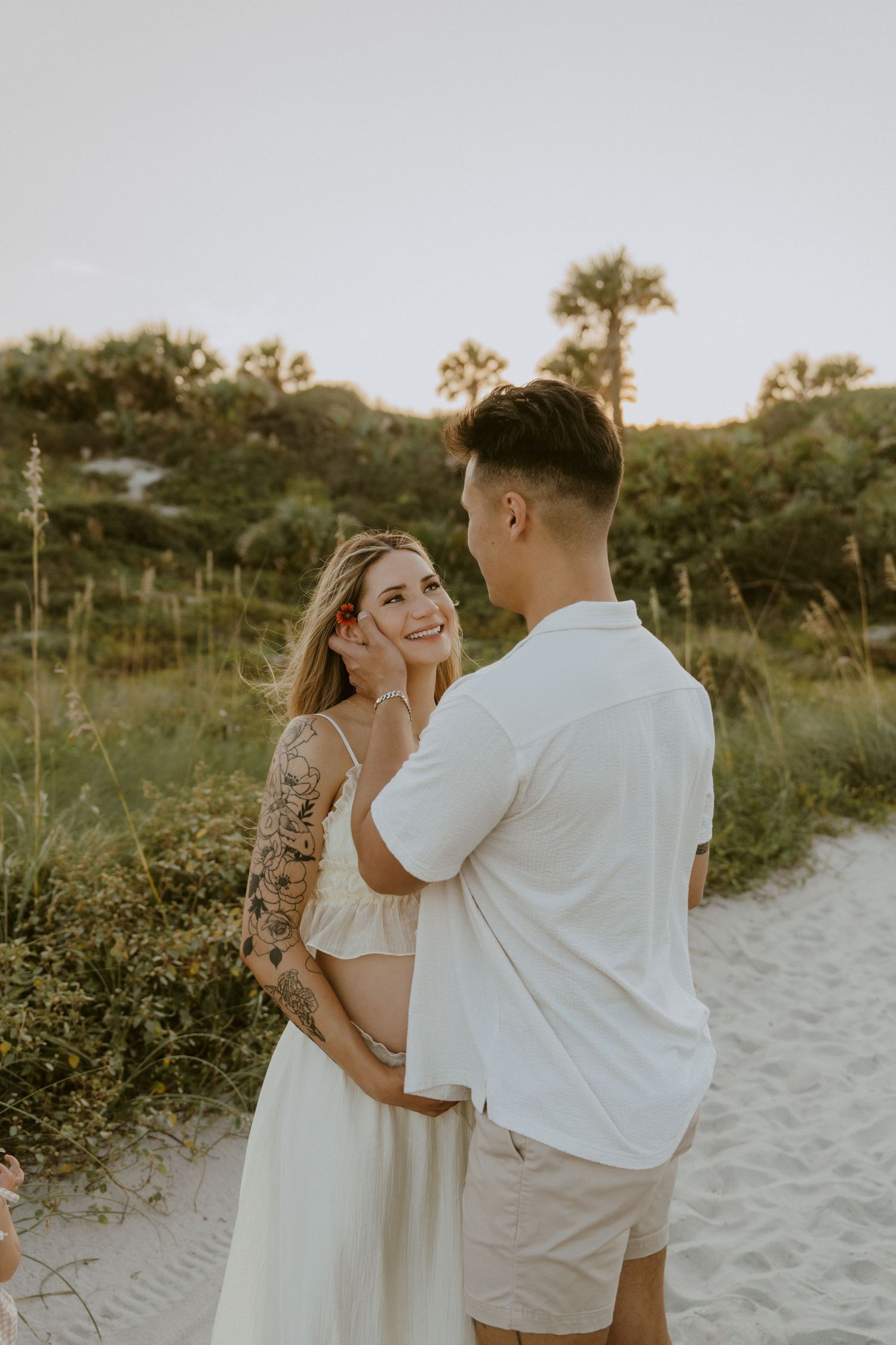 A couple in white attire walks along a sandy beach path at sunset with palm trees in the background.