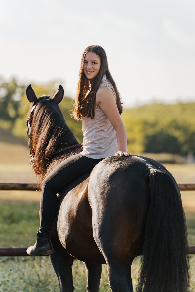 A rider sits bareback on a dark horse in a sunlit field during golden hour, creating a peaceful outdoor scene.