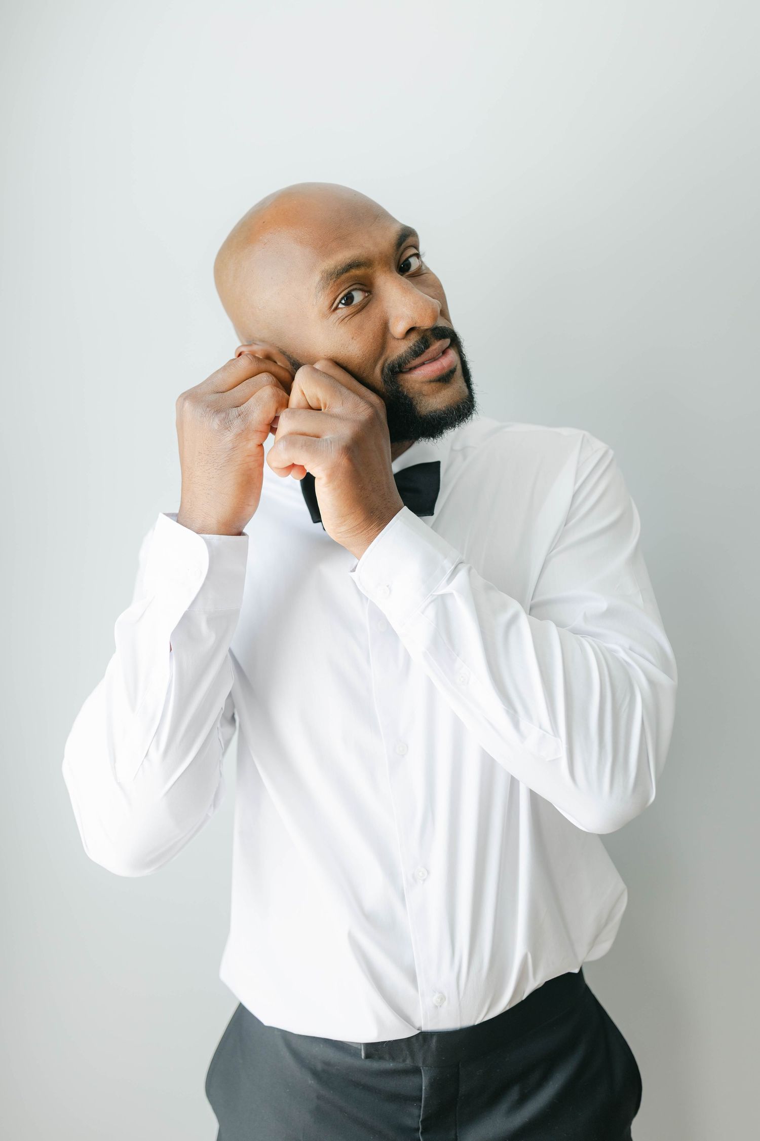A person in formal attire with a white dress shirt and black bow tie adjusts their collar against a light background.
