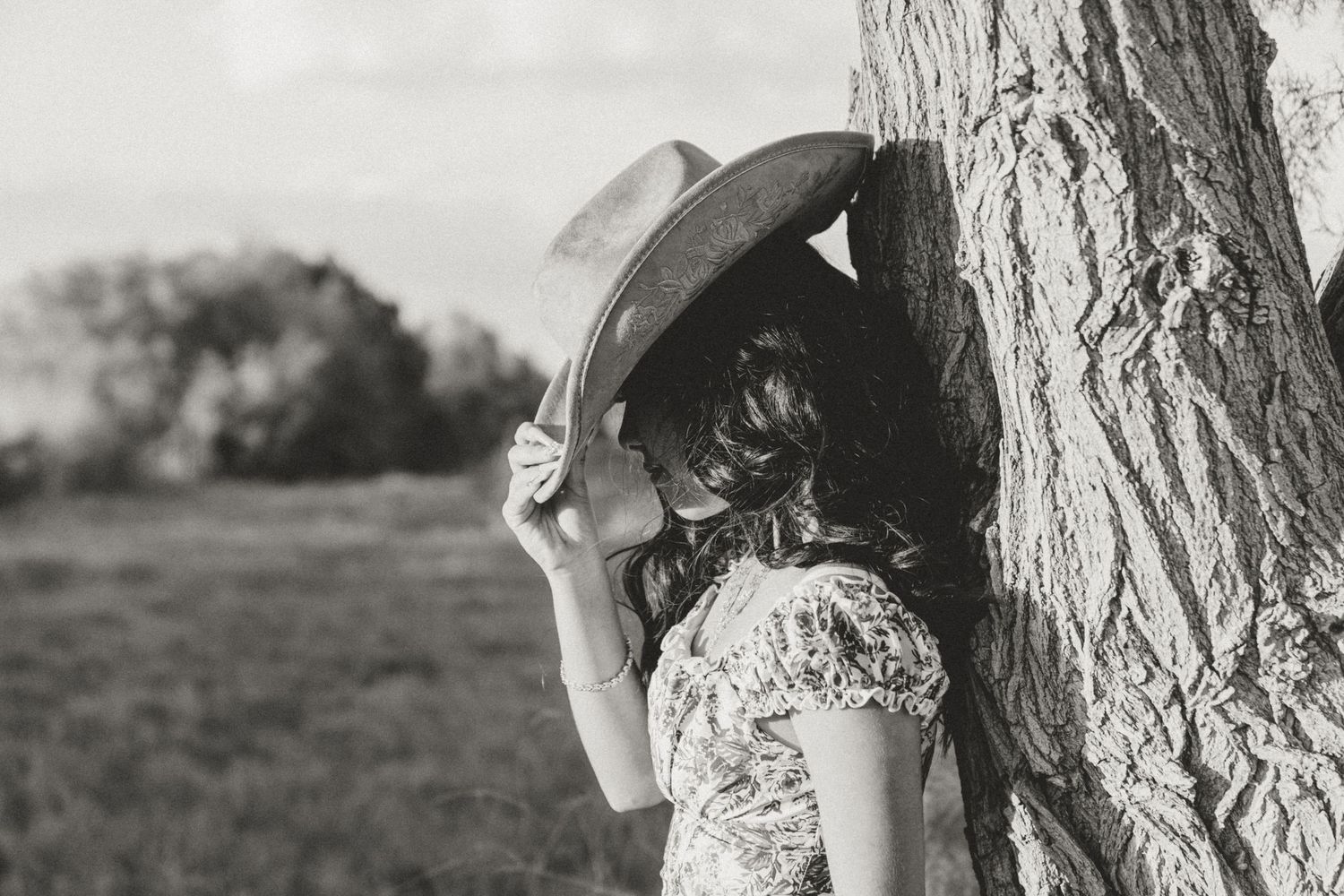 A young girl leaning on a tree, shades face from sun with cowboy hat.