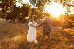 Romantic couple dancing together in a golden wheat field at sunset with warm backlighting through trees.
