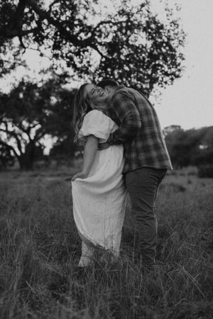 Black and white photo of a romantic embrace in a rustic field with trees silhouetted in background.