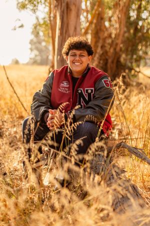 Person in varsity jacket sitting in golden field with eucalyptus trees in background.