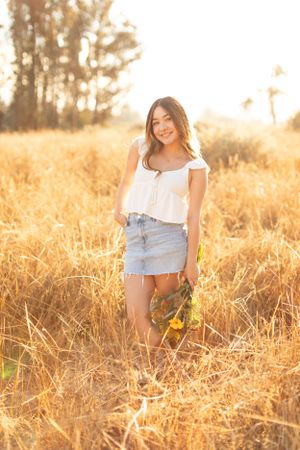 Woman in white top and denim skirt standing in golden wheat field at sunset.