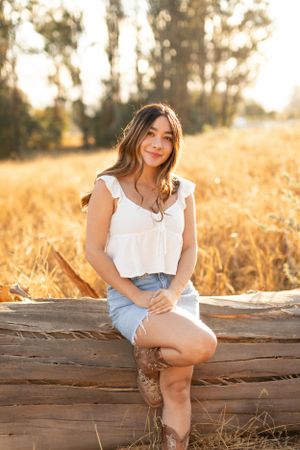 Woman in white top and denim shorts sits on rustic log in golden field at sunset.