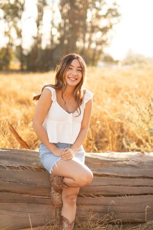 Woman posing casually on wooden log in sunlit field wearing summer outfit.