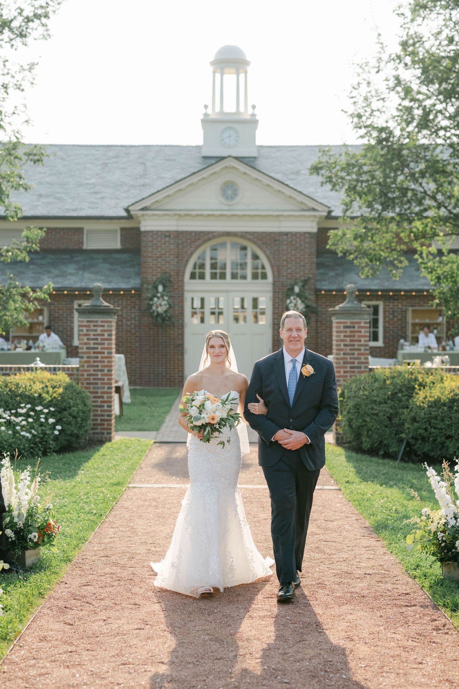 A wedding procession walking down a brick-lined path leading to a white church with arched windows and a steeple. - Elawa Farm