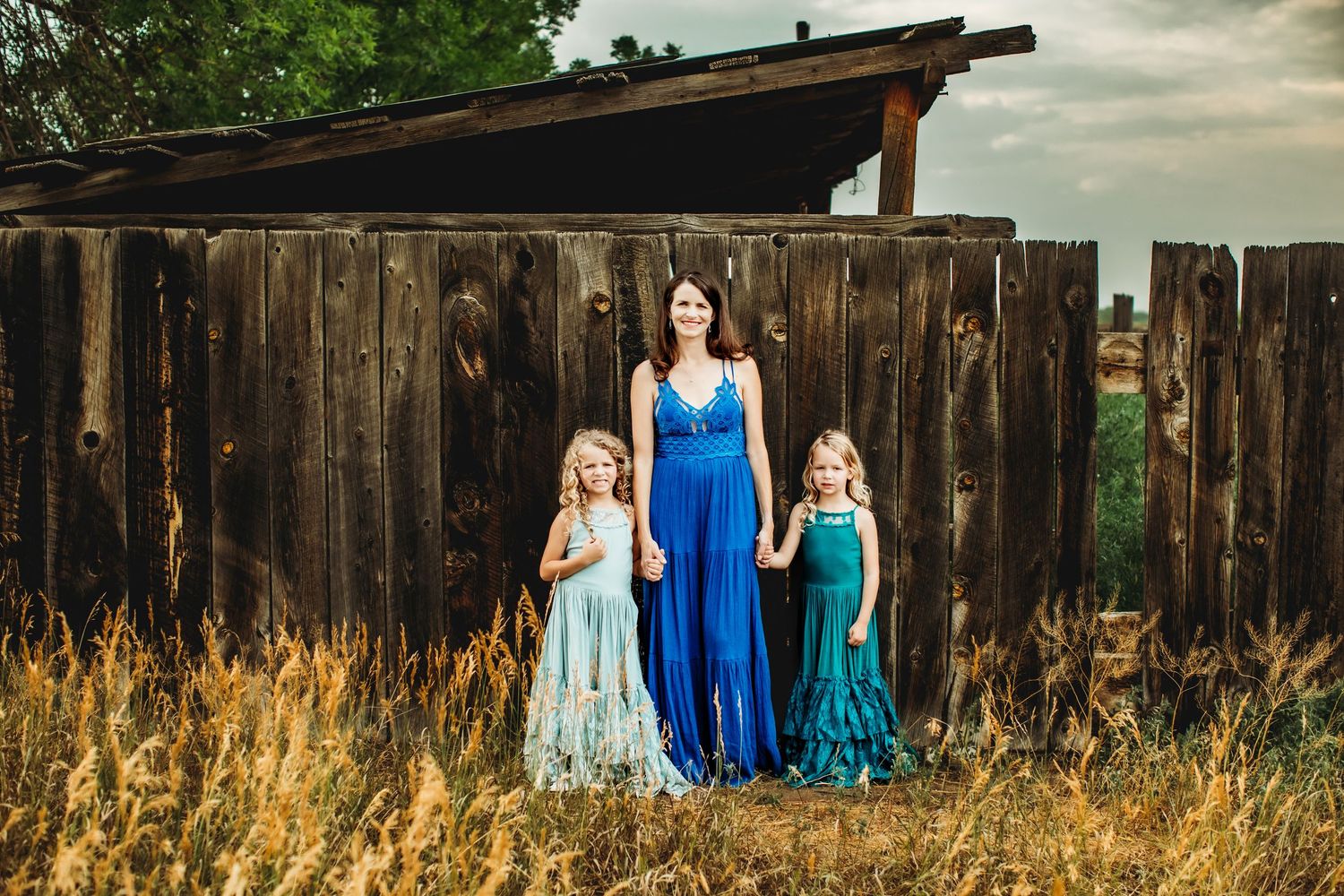 Three people in flowing blue dresses stand in front of a rustic wooden fence and barn at sunset.