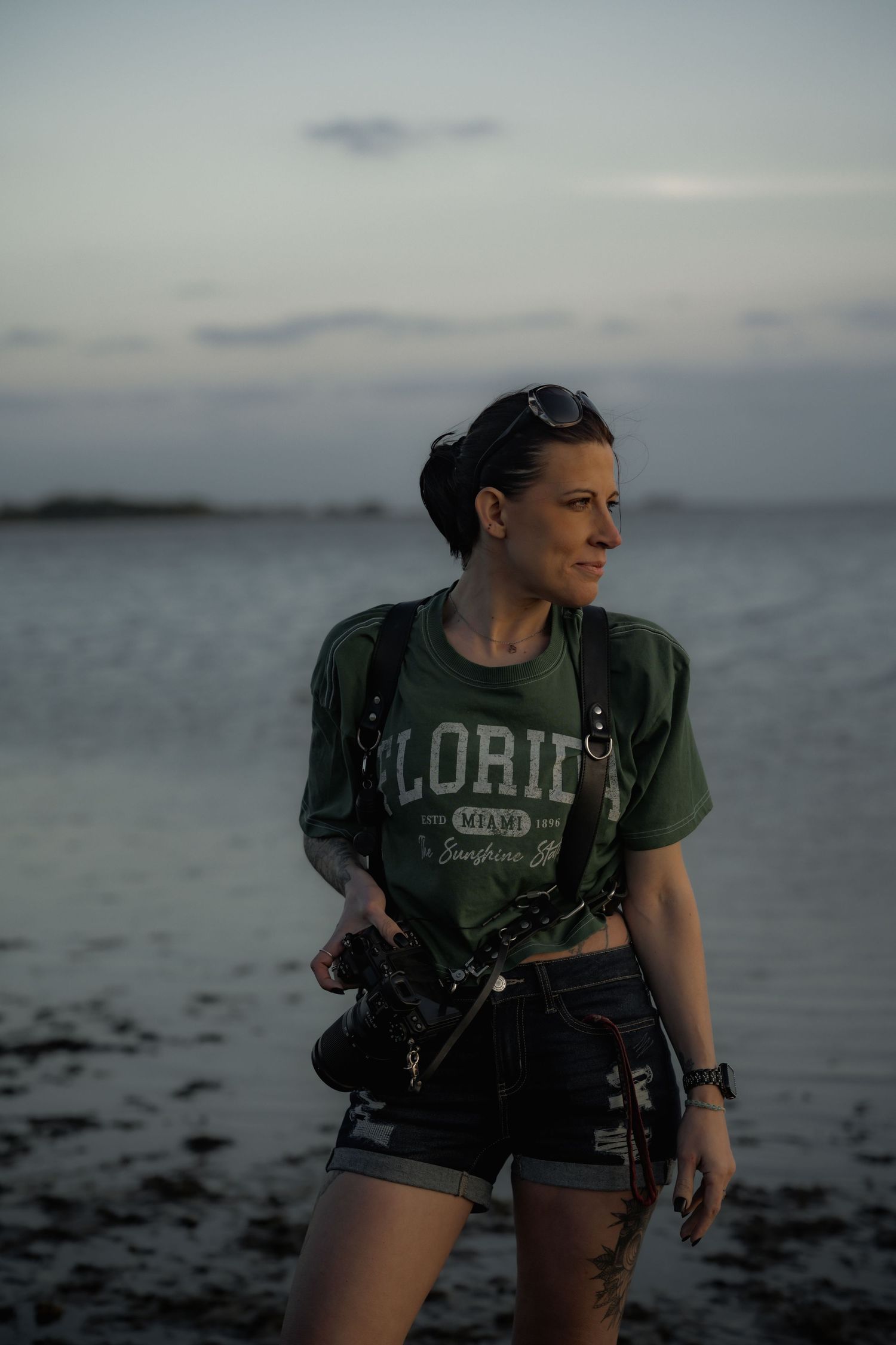 A photographer in a Florida t-shirt and denim shorts stands on a moody beach at dusk holding camera equipment.