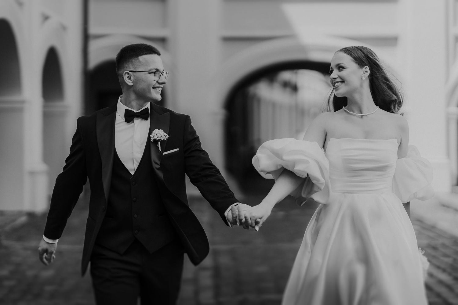 A joyful black and white wedding photo of a couple running and laughing together in formal attire.