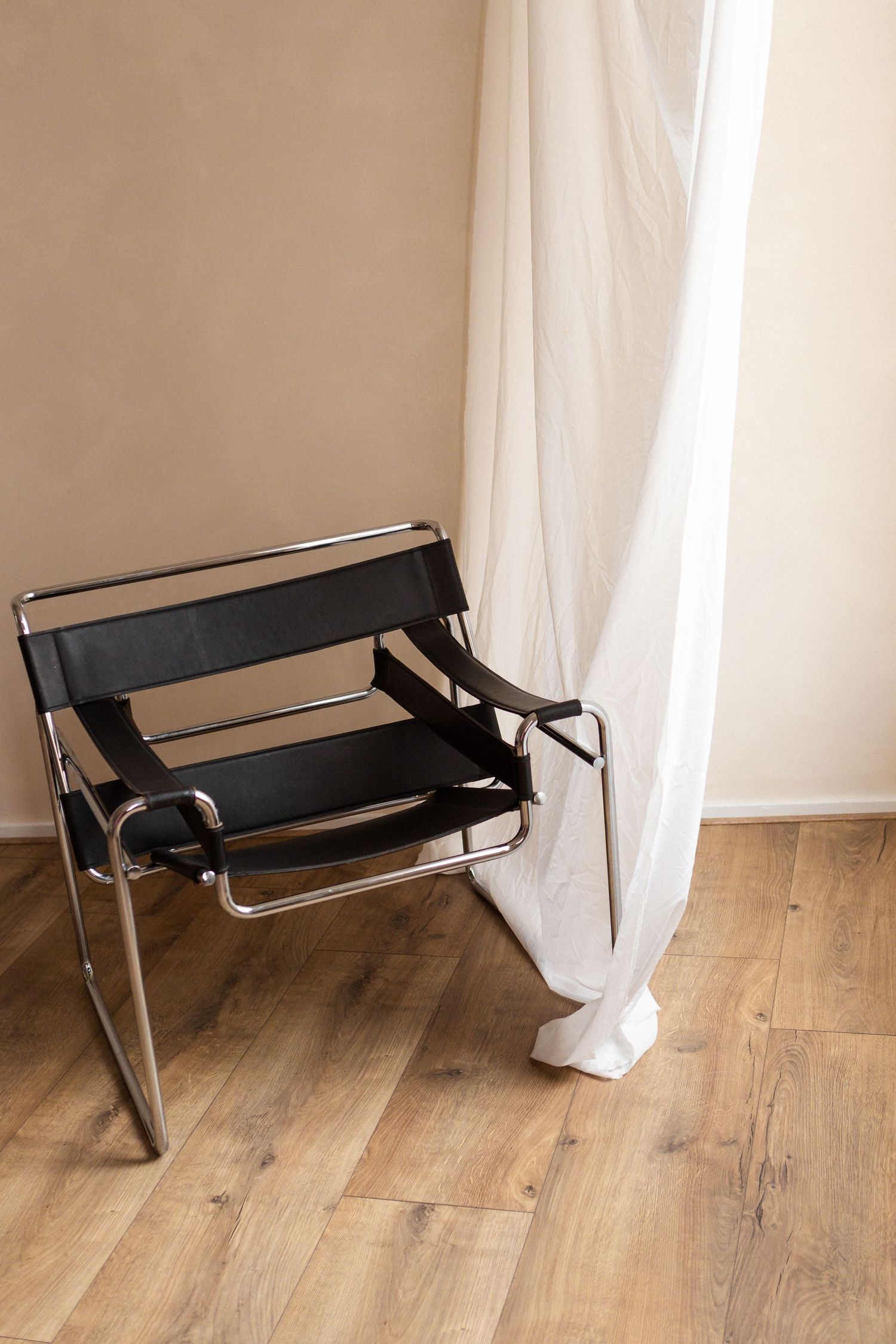 Black leather and chrome Wassily chair shown from four different angles on hardwood flooring against white curtains.