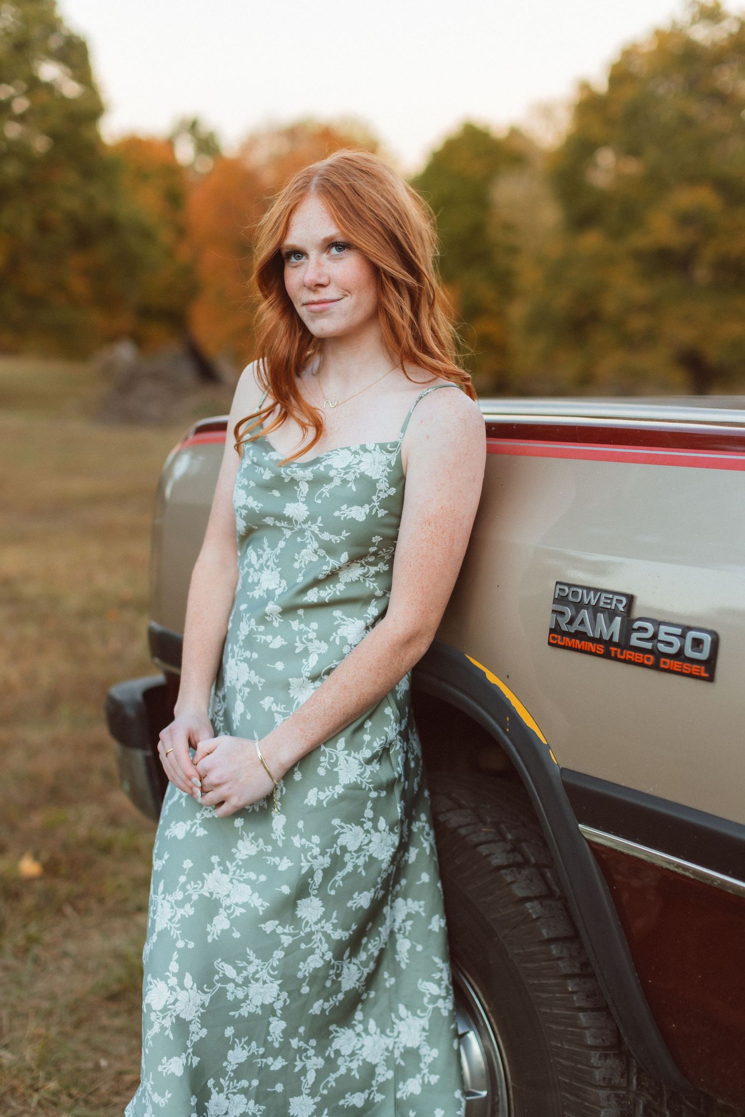 A red-headed young woman wearing a green floral dress leans against a vintage truck and smiles softly during her senior photo session.