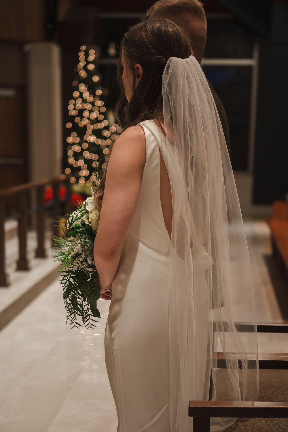 Bride in white dress with flowing veil standing at end of church aisle with blurry lighted christmas tree.