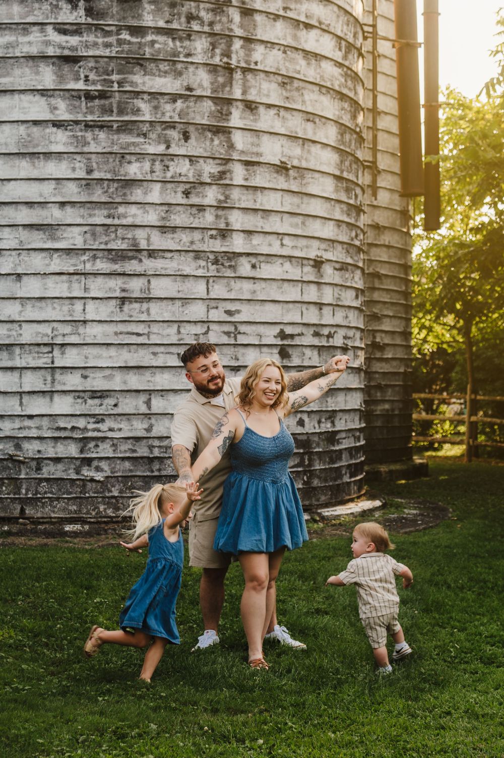 A joyful family plays and dances together in front of a rustic silo wall during golden hour.