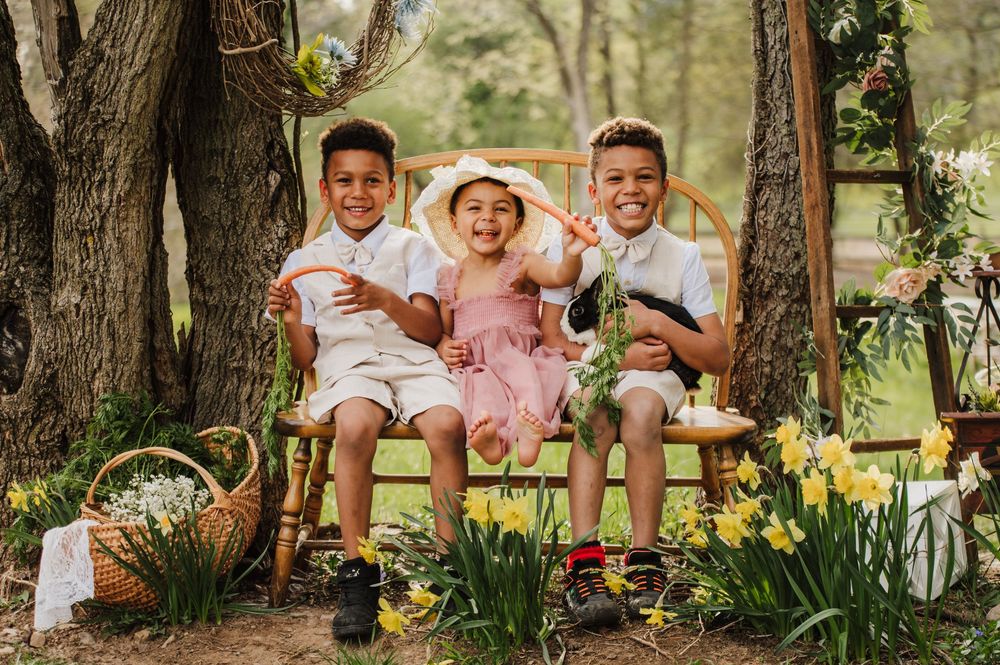 Three children sit together on a wooden swing in a garden surrounded by spring daffodils and trees.